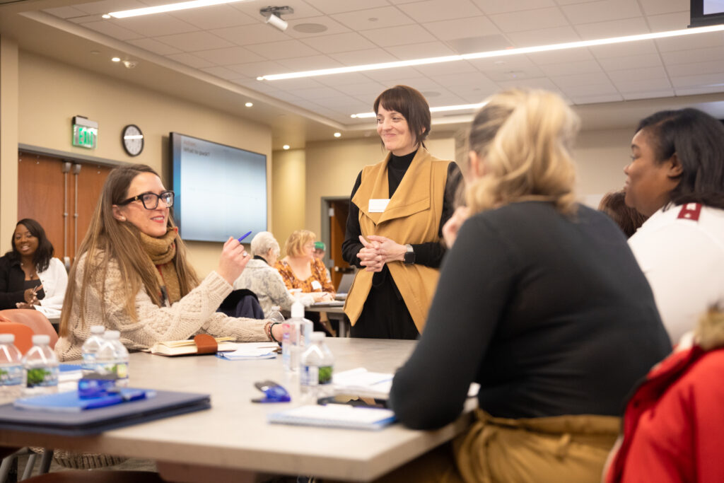 Mandy Sharp Eizinger stands at a table, speaking to seated event attendees.