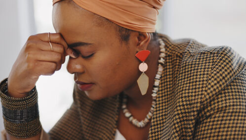 A Black woman wearing a headwrap rests her head on her hand with her eyes closed, as if stressed