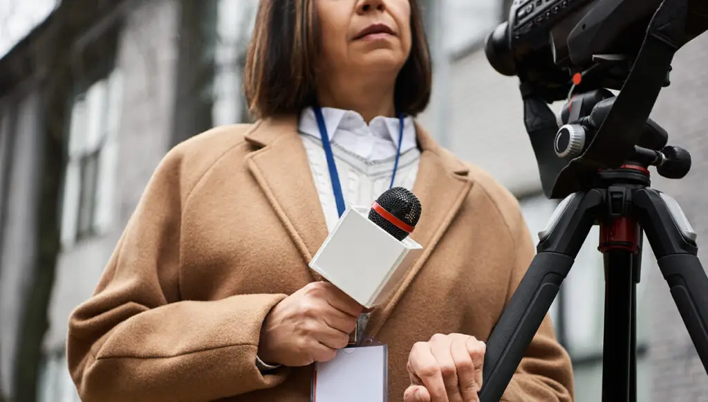 A female news journalist stands outside of a building on a gray day with a microphone and camera