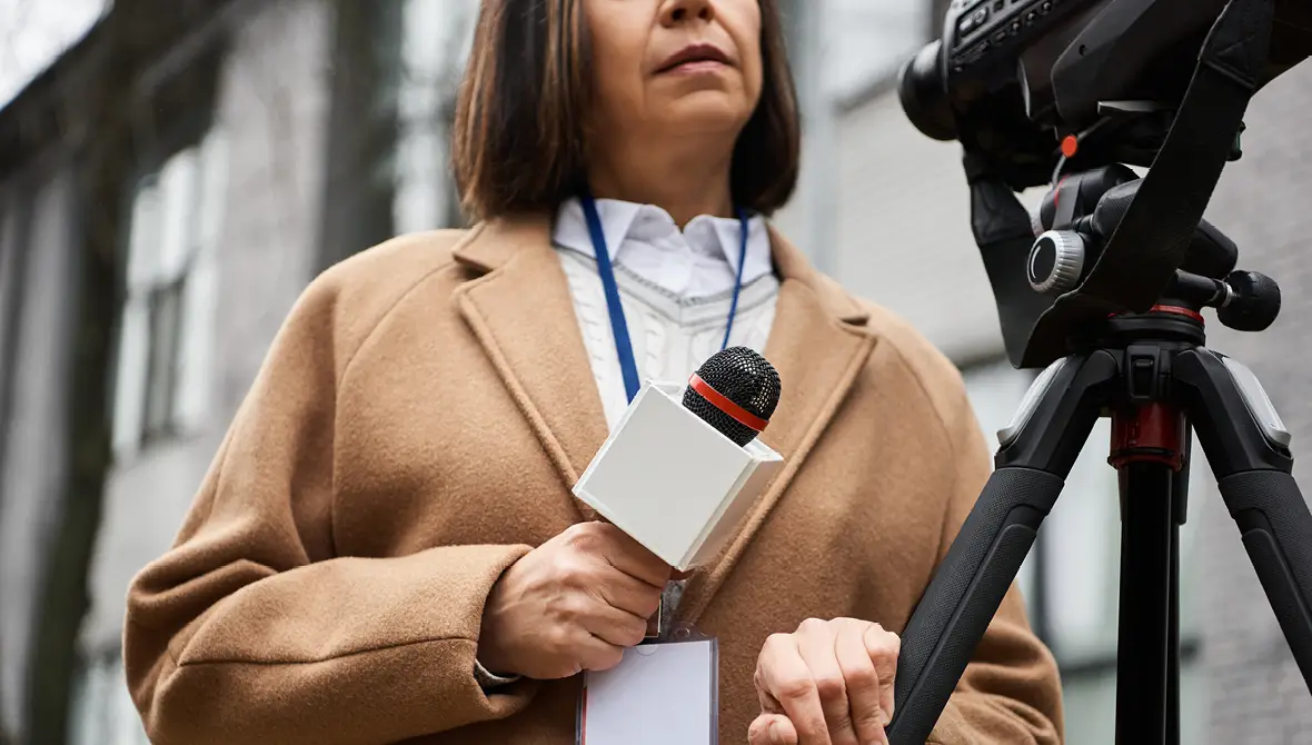 A female news journalist stands outside of a building on a gray day with a microphone and camera