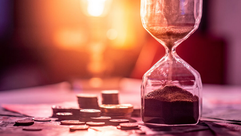 Photo of an hourglass on a table with small stacks of coins and dollar bills next to it