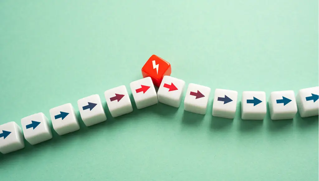 A line of white cubes with blue arrows pointing to the right is interrupted by a red cube with a lightening bolt, representing a crisis or inflection point in a lifecycle