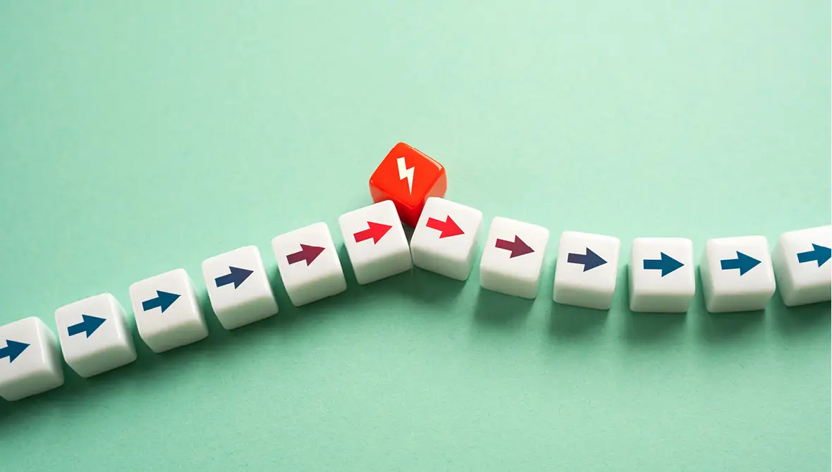 A line of white cubes with blue arrows pointing to the right is interrupted by a red cube with a lightening bolt, representing a crisis or inflection point in a lifecycle